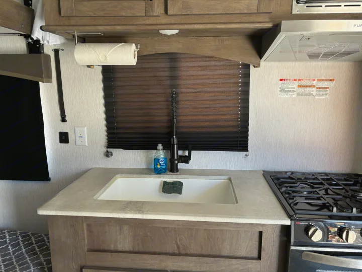 RV rental kitchen features a sink, black faucet, dish soap, and a Suburban gas stove with overhead cabinets.. Forest River Stealth 2022