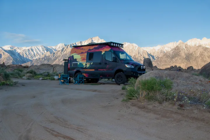 Colorful RV rental van with a custom wrap parked in a rocky desert, foregrounded by chairs and snow-capped mountains.. Mercedes-Benz Sprinter 2023