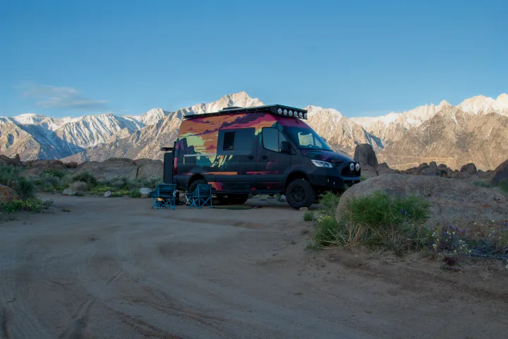 Experience off-grid camping! This colorful RV rental van is parked with chairs amidst rocks, backed by snowy mountains.. Mercedes-Benz Sprinter 2023