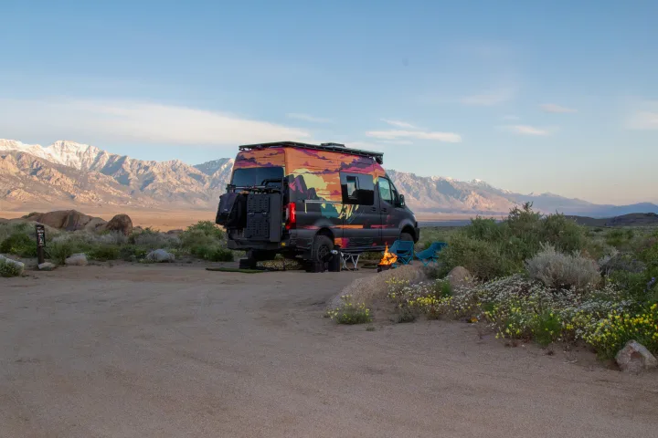 This vibrant RV rental is parked at a desert campsite, ready for adventure. Campfire glowing with stunning snow-capped mountains behind.. Mercedes-Benz Sprinter 2023