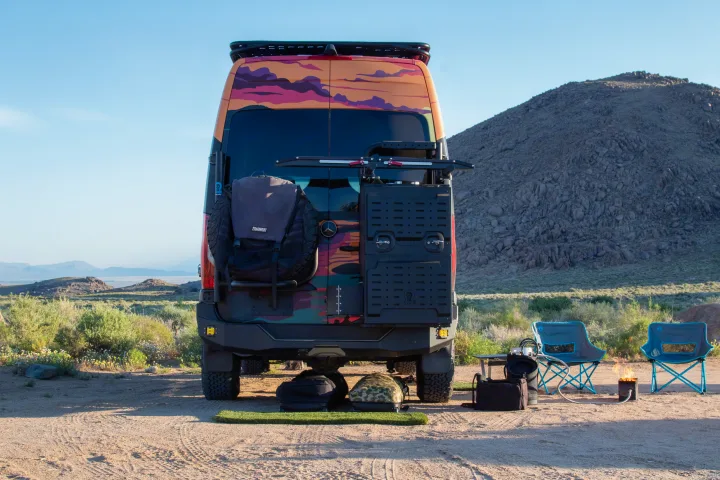 This custom RV rental is set up for desert camping, with chairs and a cozy fire pit behind the colorful van.. Mercedes-Benz Sprinter 2023