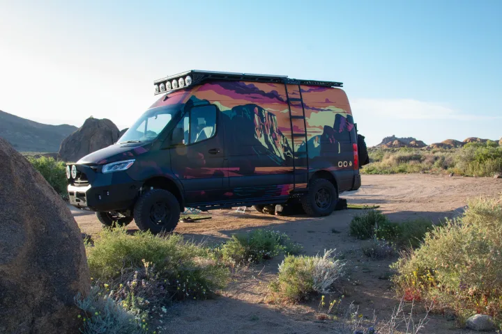 Custom RV rental parked in a desert landscape. This black adventure van features a vibrant mural, off-road tires, and roof lights.. Mercedes-Benz Sprinter 2023