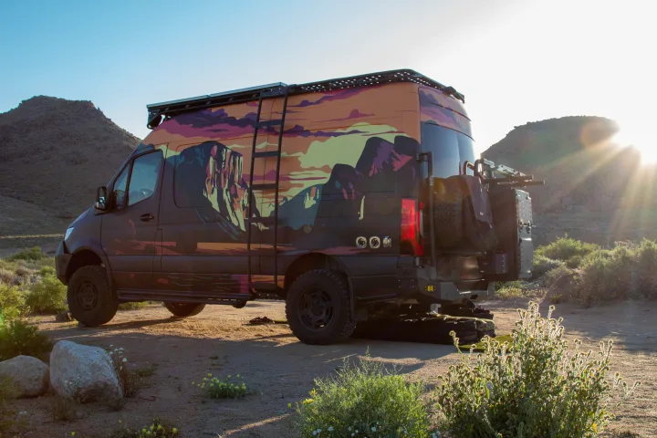 Rugged RV rental camper van, wrapped with vibrant mountain art, parked in a sunlit desert, ready for your next adventure.. Mercedes-Benz Sprinter 2023