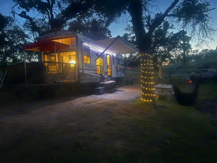 Nighttime rv rental setup: a brightly lit camper with a rear deck, string-lit tree, and picnic area for camping leisure.. Heartland 265 2021