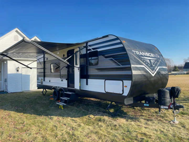 A black & white Transcend travel trailer, ready for rv rental, parked on grass beside a house with its awning out.. Grand Design Transcend Xplor 2022