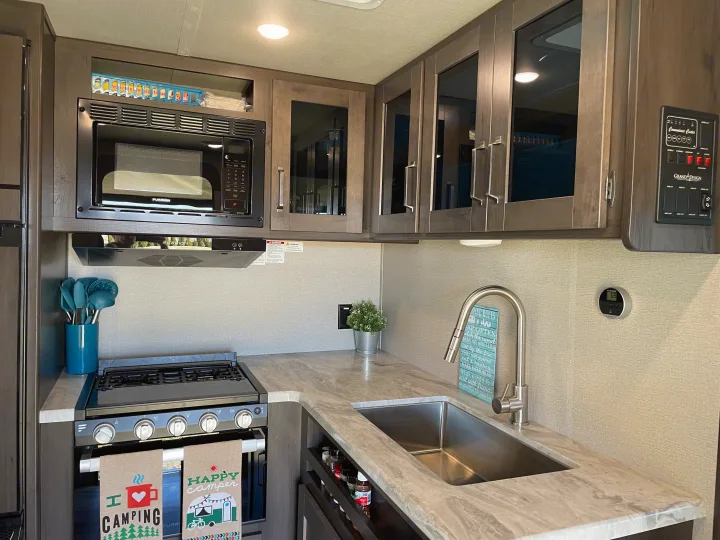 A well-equipped RV rental kitchen featuring a microwave, stove, sink, and charming camping decor. Includes modern controls.. Grand Design Transcend Xplor 2022