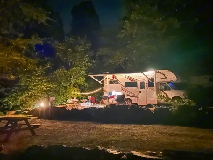 Relaxing RV rental campsite at night. Illuminated RV, picnic tables, chairs, fire pit, and string lights surrounded by trees.. Thor Motor Coach Four Winds 2019