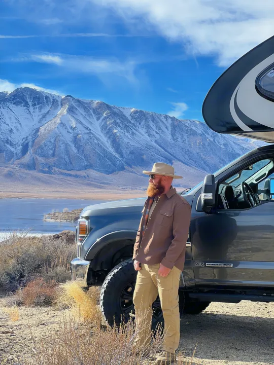 Man by RV rental truck with snowy mountains, partially frozen lake. Perfect for rugged outdoor adventures!. Ford F350 Diesel Double Cab 2020