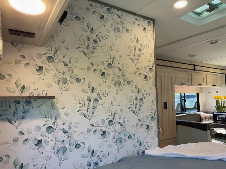 RV rental bedroom with stylish eucalyptus leaf wallpaper and cozy bed, opening to a bright living area.. Forest River Sunseeker LE 2023