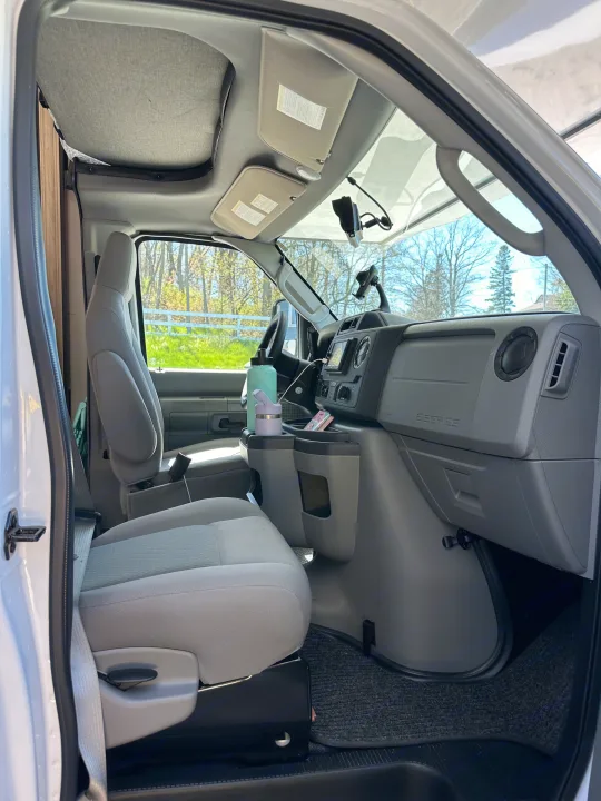 View from the side door into an RV rental's front cabin. Shows gray seats, dashboard, and custom van conversion elements.. Forest River Sunseeker LE 2023