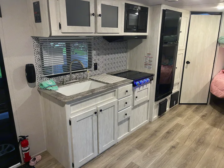 Bright RV rental kitchen with light wood cabinets, modern appliances, sink, and stove, plus a fridge & bunk area.. Shasta RVs I5 526DB 2024