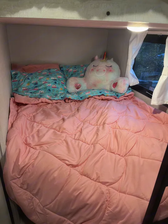 Cozy RV rental bunk bed with pink comforter, camping-themed pillow, and unicorn plush, perfect for a comfy night's sleep.. Shasta RVs I5 526DB 2024