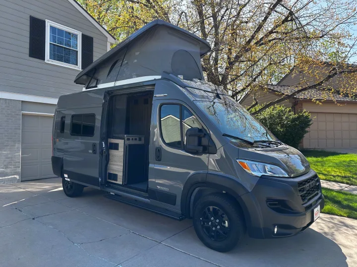 Grey pop-top camper van RV rental parked in a driveway. Side door open reveals interior. House and trees in background.. Winnebago Solis 2023