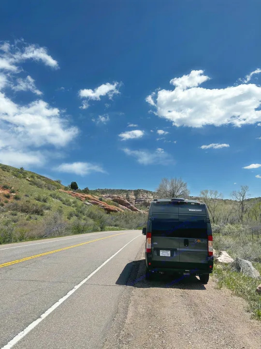 RV rental: A dark grey Winnebago Solis van parked roadside in rocky, green mountains under a bright blue, cloudy sky.. Winnebago Solis 2023