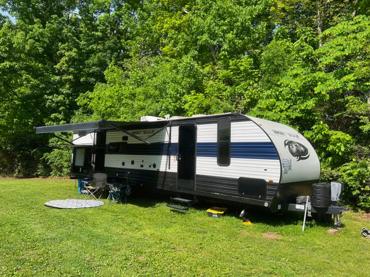 Relaxing RV rental travel trailer parked at a sunny campsite with extended awning, chairs, and mat on the green grass.. Forest River Cherokee Grey Wolf 2023