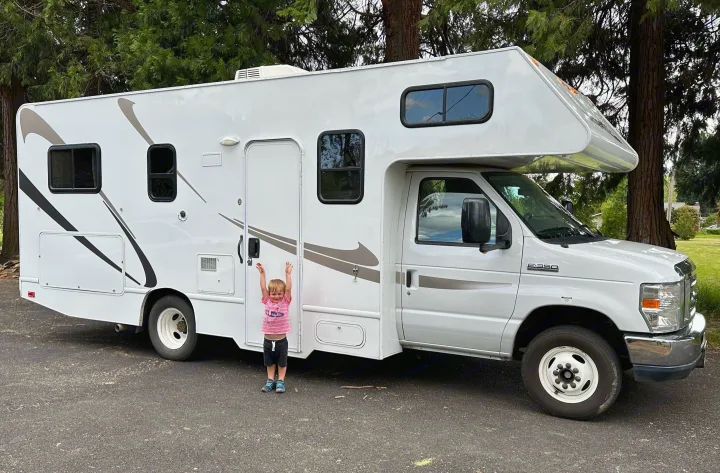 A happy child with arms raised excitedly stands beside a white RV rental, perfect for family adventures.. Thor Majestic 2015