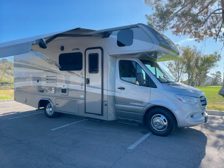 Modern silver RV rental parked on asphalt with awning extended, ready for an adventure amidst scenic hills and trees.. Forest River Dynamax Isata 3 24FW 2023