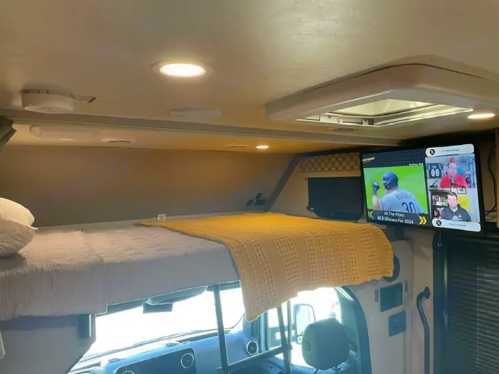 This RV rental interior features a comfortable bunk bed with a yellow blanket and a TV showing sports, ideal for travel.. Forest River Dynamax Isata 3 24FW 2023
