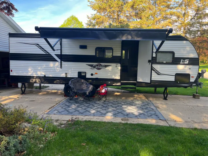 This Shasta RV rental is set up for outdoor living with an extended awning, grey mat, and two chairs on a paved driveway.. Shasta RVs I5 526DB 2024
