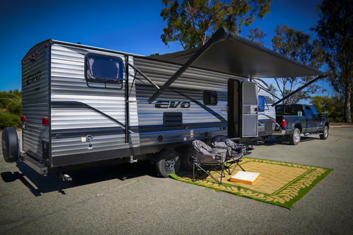 RV rental setup: a travel trailer with an extended awning, two chairs, and a mat on the ground, hitched to a pickup truck.. Forest River EVO 2021