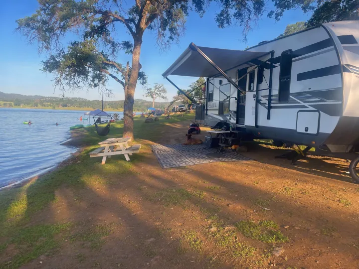 Spacious RV rental parked lakeside with awning out, a relaxing dog, picnic area, and people enjoying water activities.. Heartland 265 2021