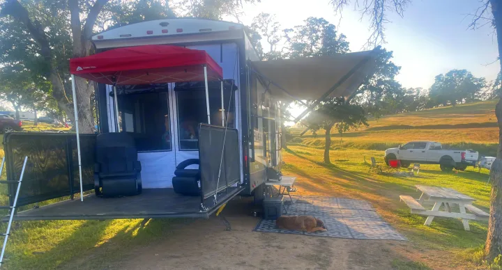 Relaxing RV rental setup! A large RV with its back deck open, covered by a red canopy, is parked in a grassy campsite with a dog napping.. Heartland 265 2021