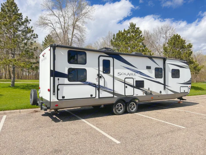 White Solaire RV rental travel trailer parked in a lot, ready for camping. Features multiple windows and a spare tire.. Forest River Solaire 2020