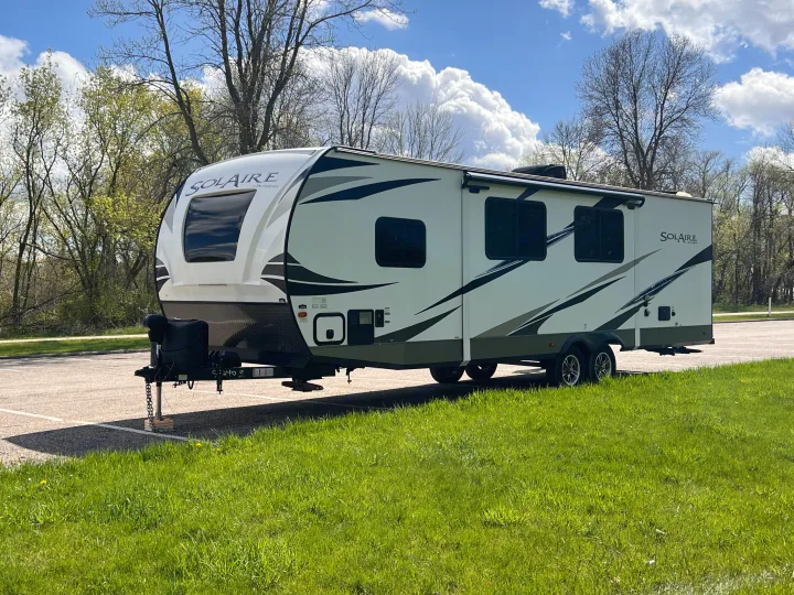 Bright white Solaire travel trailer, ideal for RV rental, parked on asphalt with green grass & trees under a blue sky.. Forest River Solaire 2020