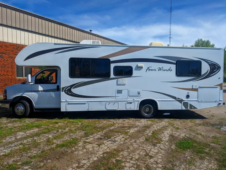 White Class C RV rental, a Four Winds motorhome with brown & gray decals, parked on a gravel lot under blue sky.. Thor Motor Coach Four Winds 2014