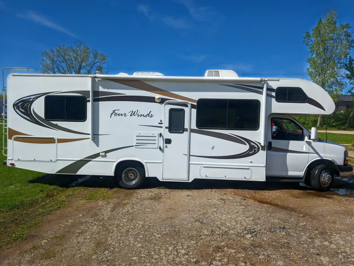 White Four Winds RV rental, a clean motorhome with brown trim, parked on gravel with grass under a clear blue sky.. Thor Motor Coach Four Winds 2014