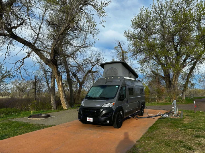 RV rental camper van with pop-top at a spring campsite, hooked up to utilities by a fire pit.. Winnebago Solis 2023