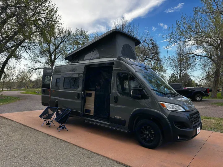 A gray Winnebago Solis camper van RV rental, with its pop-top extended and side door open, parked for camping.. Winnebago Solis 2023