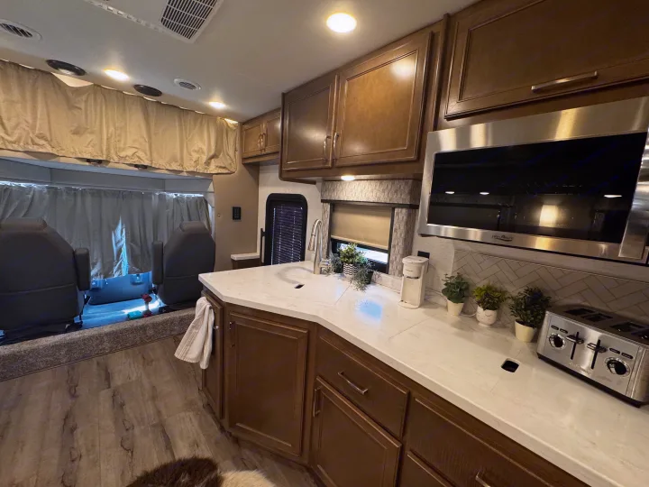 Inside an RV rental: a modern kitchen with wood cabinets, white counters, and microwave, plus driver's seats. Cozy home for travel.. Renegade Verona 40 VRB 2024