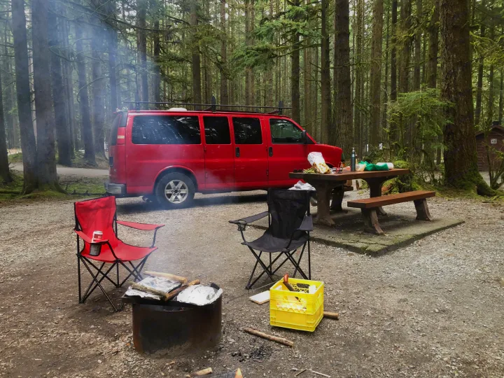 A red RV rental van at a serene forest campsite. Features a smoking fire pit, camping chairs, and a picnic table setup.. Gmc Savanna 2010