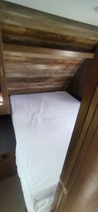 Cozy sleeping area in an RV rental. A bed with a white fitted sheet is tucked into a compact space with rustic wood-paneled walls.. Keystone RV Passport 2019