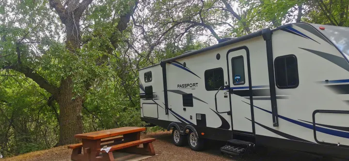 Enjoy a serene RV rental camping experience! This travel trailer is parked amidst lush trees with a picnic table nearby.. Keystone RV Passport 2019