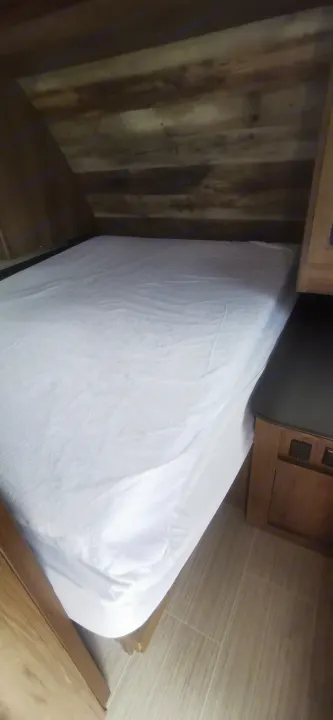 Cozy bed with white sheets in an RV rental's bedroom. Features rustic wood paneling, angled ceiling, and bedside charging outlets.. Keystone RV Passport 2019