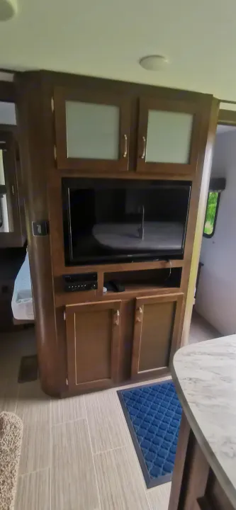 View of an RV rental interior: a wooden entertainment center with TV and cabinets, tiled floor, and a blue rug. Cozy living space.. Keystone RV Passport 2019