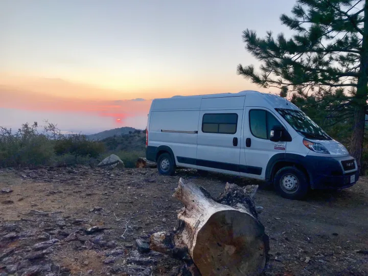 A white RV rental camper van parked on a dirt road, overlooking a vibrant mountain sunset.. ram promaster 2020
