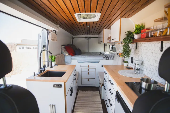 Bright RV rental interior: a modern van conversion with light wood counters, white cabinets, kitchenette, and cozy bed.. Dodge Ram Promaster 2021