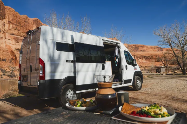 A white RV rental with an open side door is parked by a river, red rock cliffs, and bare trees, with breakfast on a table.. Dodge Ram Promaster 2021