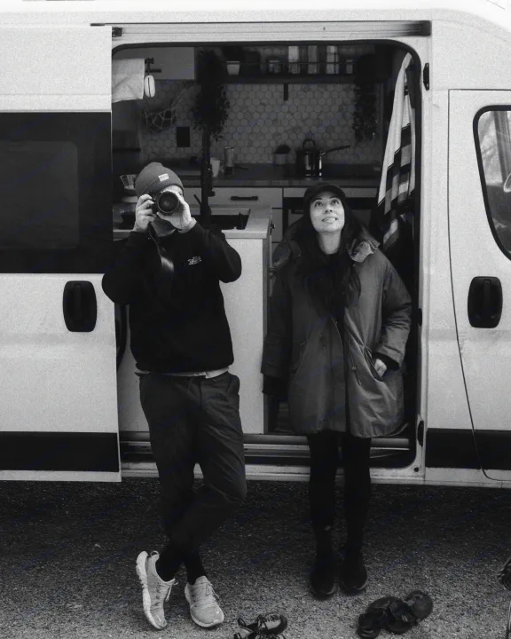 A couple by their white RV rental, she looking up, he snapping a photo. Inside, a cozy camper van kitchen awaits.. Dodge Ram Promaster 2021