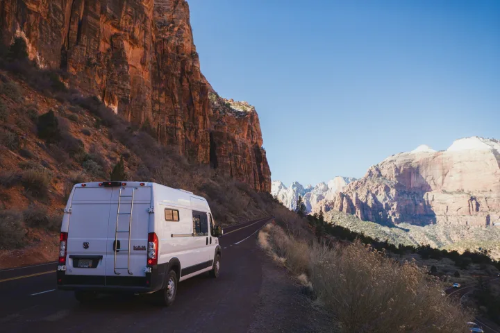 This RV rental van is parked on a winding road amidst the towering red cliffs of Zion National Park. Clear blue sky.. Dodge Ram Promaster 2021