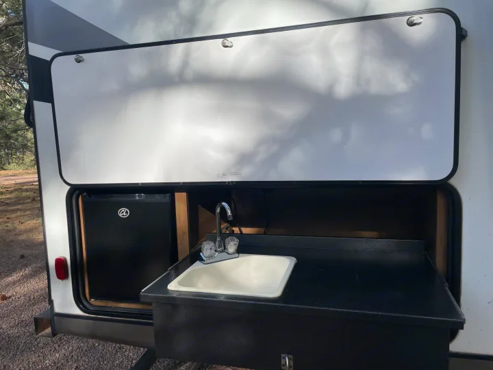 An open compartment on an RV rental reveals a practical outdoor kitchen setup with a mini-fridge, sink, and counter.. Gulf Stream Envision 2020