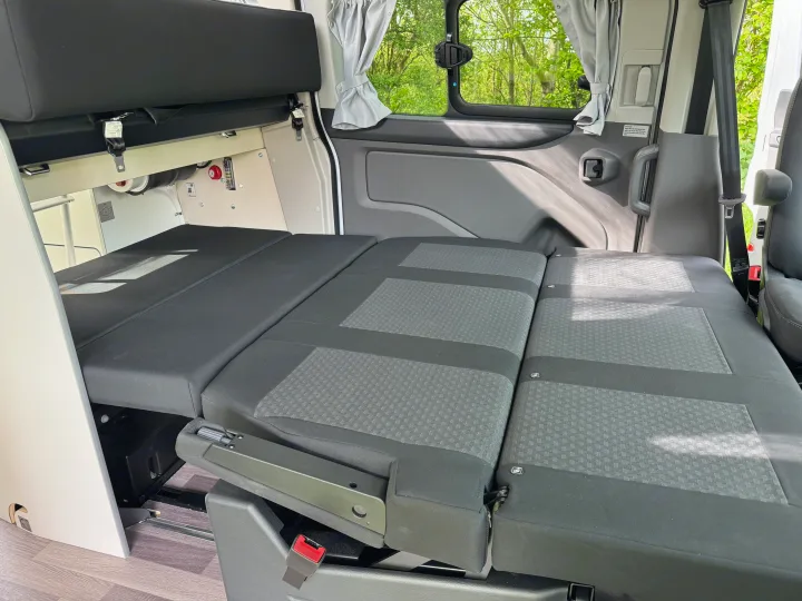 Inside an RV rental: rear seats fold down to a bed. Features windows with curtains, safety belts, and storage.. Ford Nugget 2022