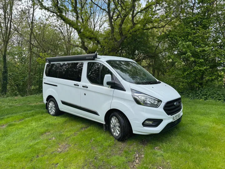 Pristine white Ford Transit Westfalia camper van on lush grass, nestled among trees. Your ideal RV rental.. Ford Nugget 2022