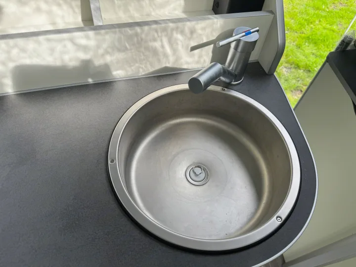Compact sink and faucet in a modern RV rental kitchen, featuring a dark counter and outdoor grass view.. Ford Nugget 2022
