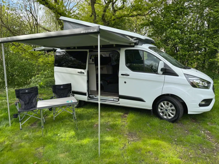 White Westfalia camper van RV rental at a green campsite with an open awning, chairs, and table set up.. Ford Nugget 2022