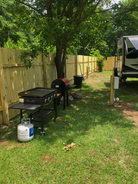 Outdoor cooking area at an RV rental site with a travel trailer, grills, and propane tank beside a wooden fence and large tree.. Grand Design Imagine 2023