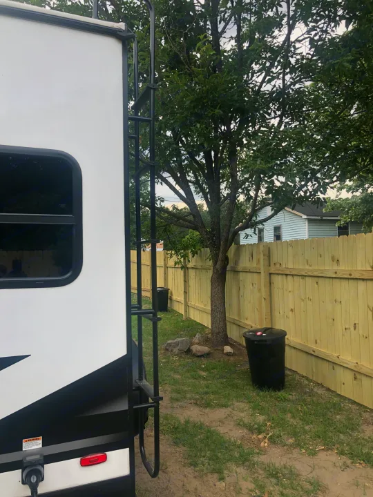 View of an RV rental's rear, showing its ladder, parked beside a new wooden fence, tree, and yard. Ideal for camping.. Grand Design Imagine 2023
